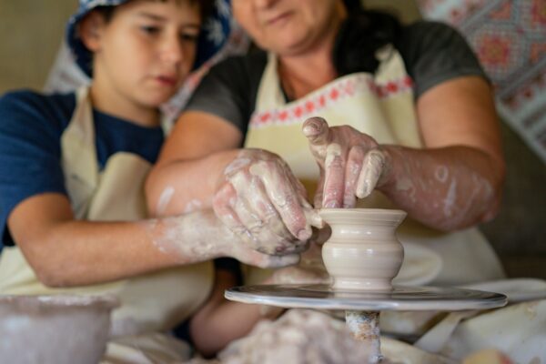Mother guiding her son in a pottery craft session, shaping clay bowl on pottery wheel.