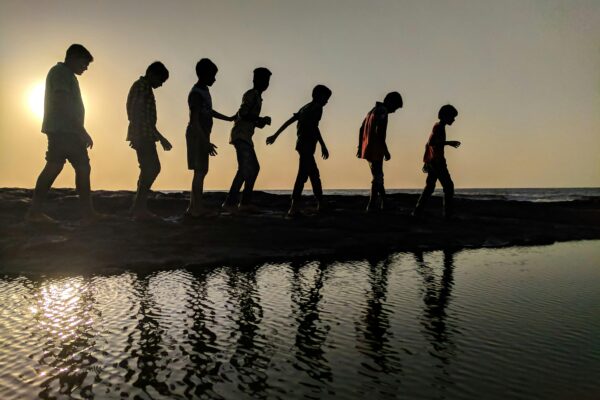 A group of children walk along the shoreline, silhouetted against a sunset, reflecting in the water.